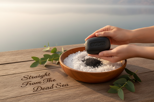 Hand holding a bar of soap above a bowl of Dead Sea salt with 'Straight From The Dead Sea' text on a wooden surface.